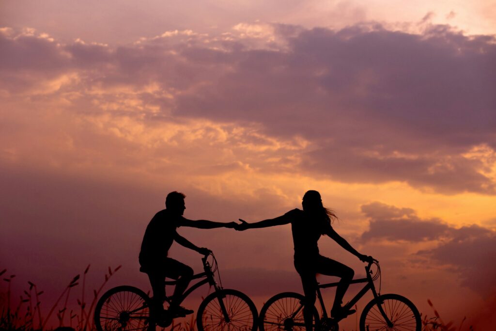 Image of a couple riding individual bicycles, holding hands, on a beautiful road with the sun setting behind them.