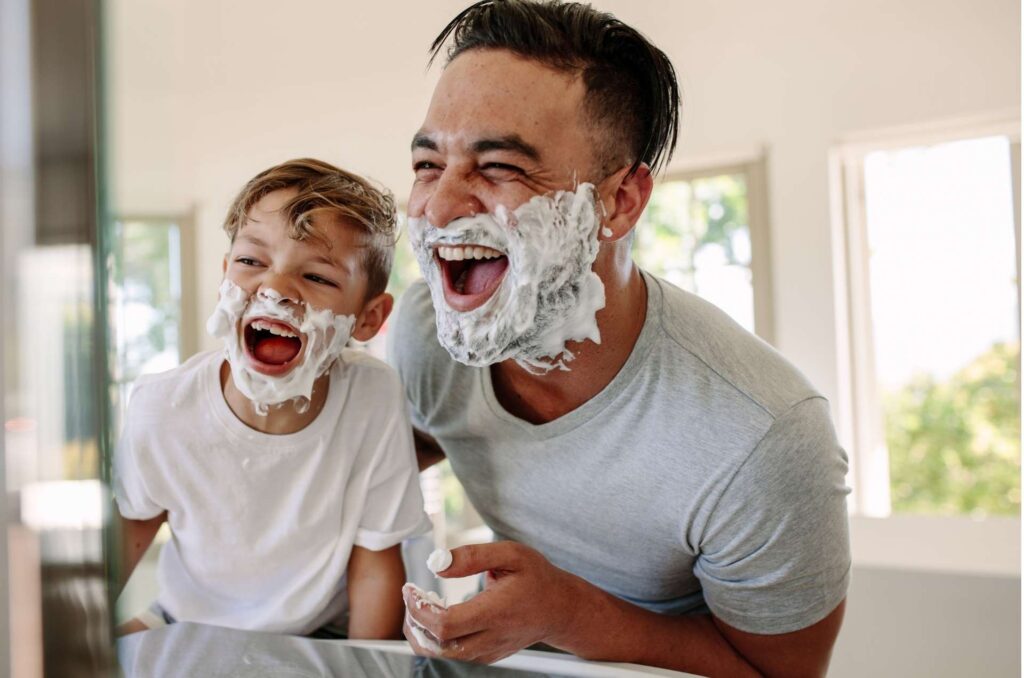 A father and son are laughing in front of a mirror with shaving cream on their faces