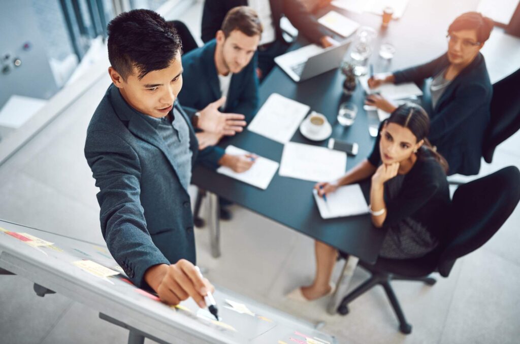 A man with a marker stands in front of a board while men and women look at the work intently.