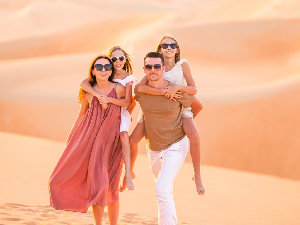 A smiling man and woman are giving two girls a piggyback ride in the desert. 