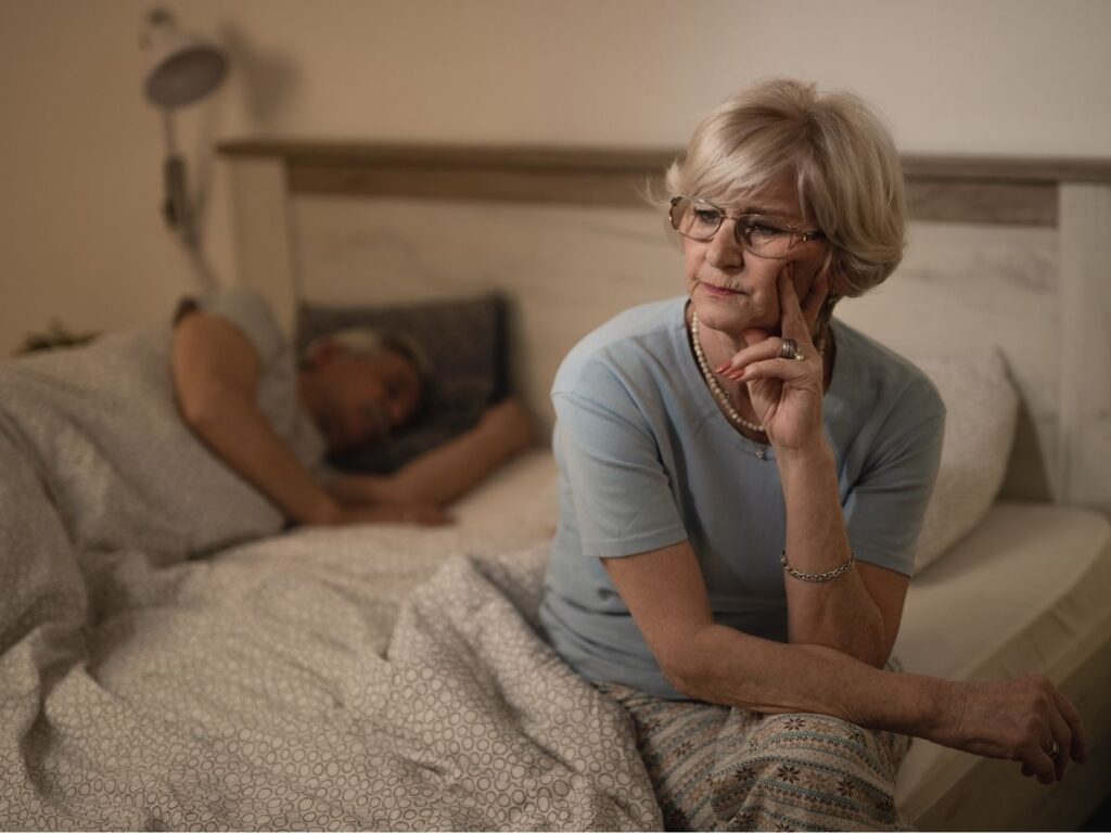 A woman sits on a bed while a man sleeps beside her